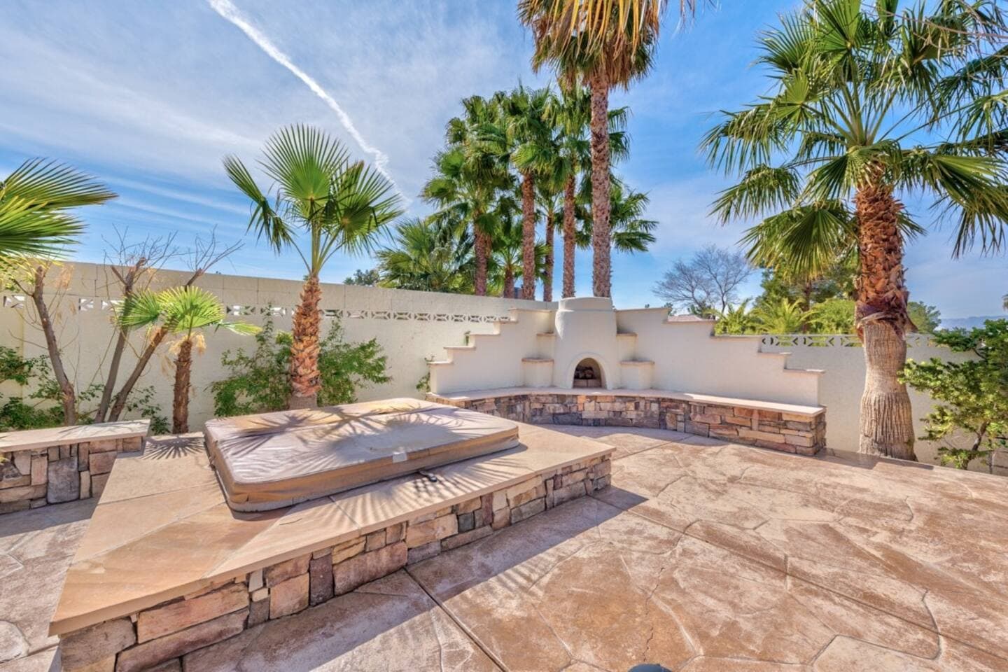  Escape to this serene stone patio featuring a raised, tiled hot tub with a cover. The area is accented by a custom, built-in outdoor fireplace and stepped seating, all surrounded by lush palm trees, 